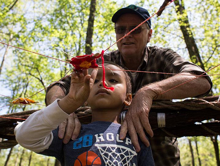 Caleb Fredrickson, 7, of Carbondale, looks at an eye of the world he tied onto a dreamcatcher with the assistance of Randy Osborn, the executive director of the Boys and Girls Club of Carbondale, Saturday, April 23, 2016, at Touch of Nature Environmental Center. The club, which partnered with Carbondale Community Arts, received a grant from the National Endowment for the Arts to implement community art projects, Osborn said. Since then, seven community art projects have been completed through the Here We Live: Arts Build Community series. One of the goals of the project was to beat the current world record holder for the largest dreamcatcher, which according to Guinness World Records has a diameter of 9 feet, 10 inches. The dreamcatcher constructed for Saturday has a diameter of 14 feet. We wanted people to think about the dreams they have, an aspiration, said Osborn, referring to why participants tied eyes of the world onto the dreamcatcher. (Aidan Osborne | Daily Egyptian)