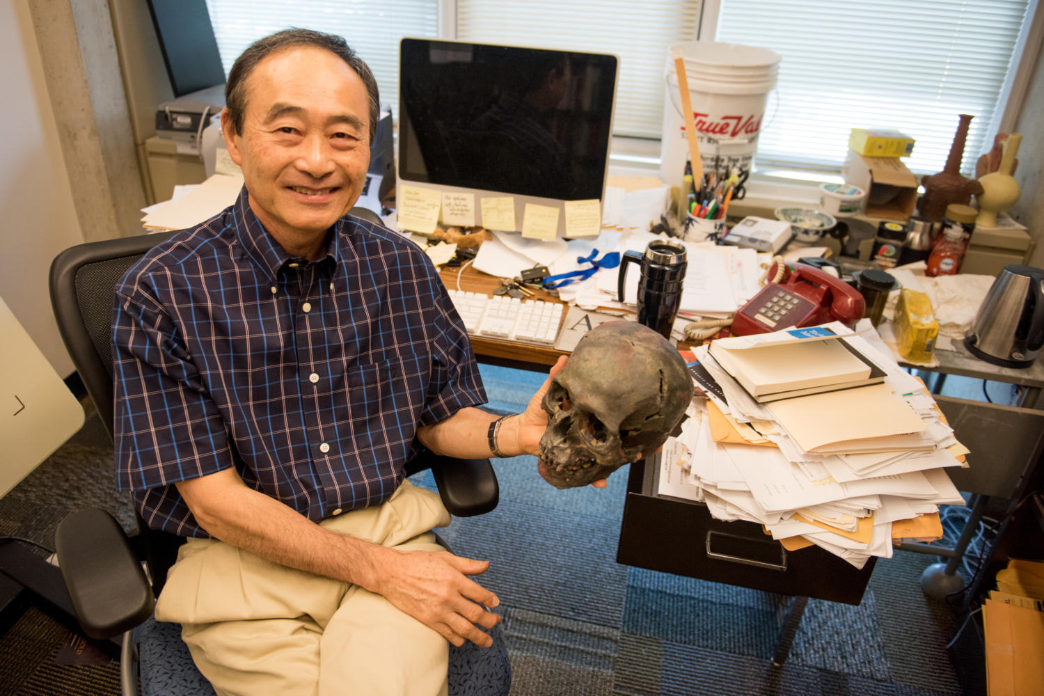 Professor of Anthropology Izumi Shimada, of Carbondale, flashes a smile for a portrait Monday, Aug. 28, 2017, at his office in Faner Hall. Shimada has been studying the Peruvian subculture, which he named the Sicans, for the past 38 years. “You have to learn how to take care of human remains,” Shimada said. The skull that Shimada is holding is a replica of a skull found while excavating in Peru that was 3-D printed at the Tokyo Metro Police Headquarters in Tokyo, Japan. (Brian Muñoz | @BrianMMunoz)