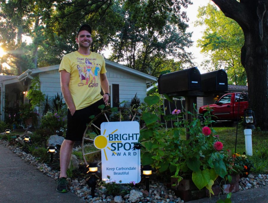 Rick+Crossley+stands+in+front+of+his+yard+beside+his+Bright+Spot+award+from+Keep+Carbondale+Beautiful+Aug.+12%2C+2021+in+Carbondale%2C+Ill.+.+Since+winning+the+award%2C+he+has+witnessed+an+increase+in+members+of+the+community+stopping+by+and+connecting+with+each+other.+%E2%80%9CIt+touches+my+heart+when+people+I+know+did+not+have+any+interactions+come+and+I+see+them+walking+by+together+in+the+mornings.+It%E2%80%99s+really+touching%2C+%E2%80%9D+Crossley+said.