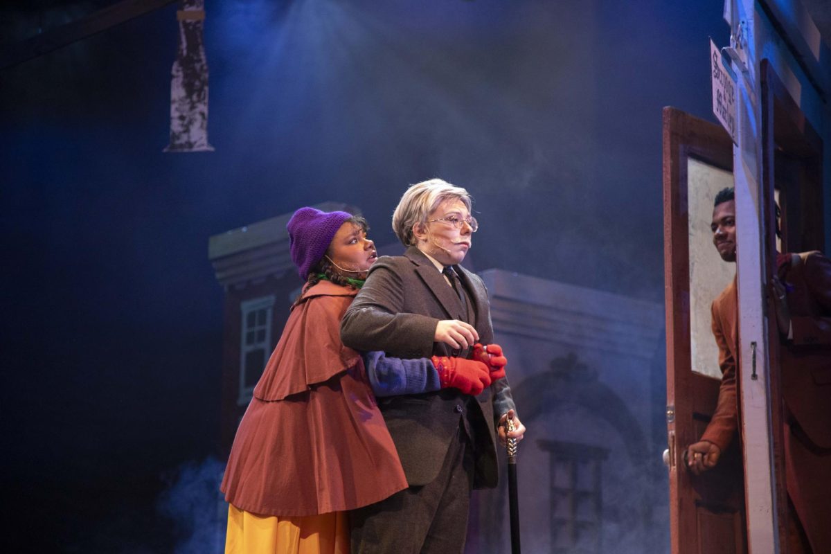 Belinda Cratchit (Yasmin Martinez-Powell) hugs an unhappy Ebenezer Scrooge (Sawyer Richman) from behind as Bob Cratchit (J’kuion West) peaks through a door Dec. 4, 2024 at McLeod Theater in Carbondale, Illinois. “I really like the end. I don't say a lick at the end but that's not why it's my favorite…I get to hug Scrooge again… so I think that's a really fun full circle moment,” Martinez-Powell said. 